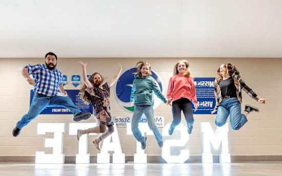 Five students jumping for joy over a 大, light-up M州 sign